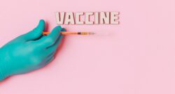 Photo of the word Vaccines with a hand wearing green surgical gloves holding a syringe below it. All on a pink background.