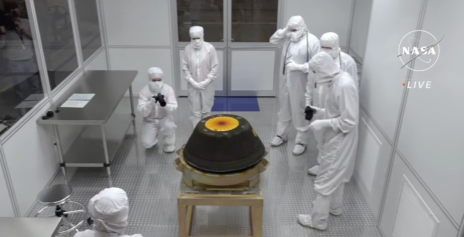 OSIRIS REx capsule in the clean room at Utah Test and Training Range. It is surrounded by 6 people in white coveralls.