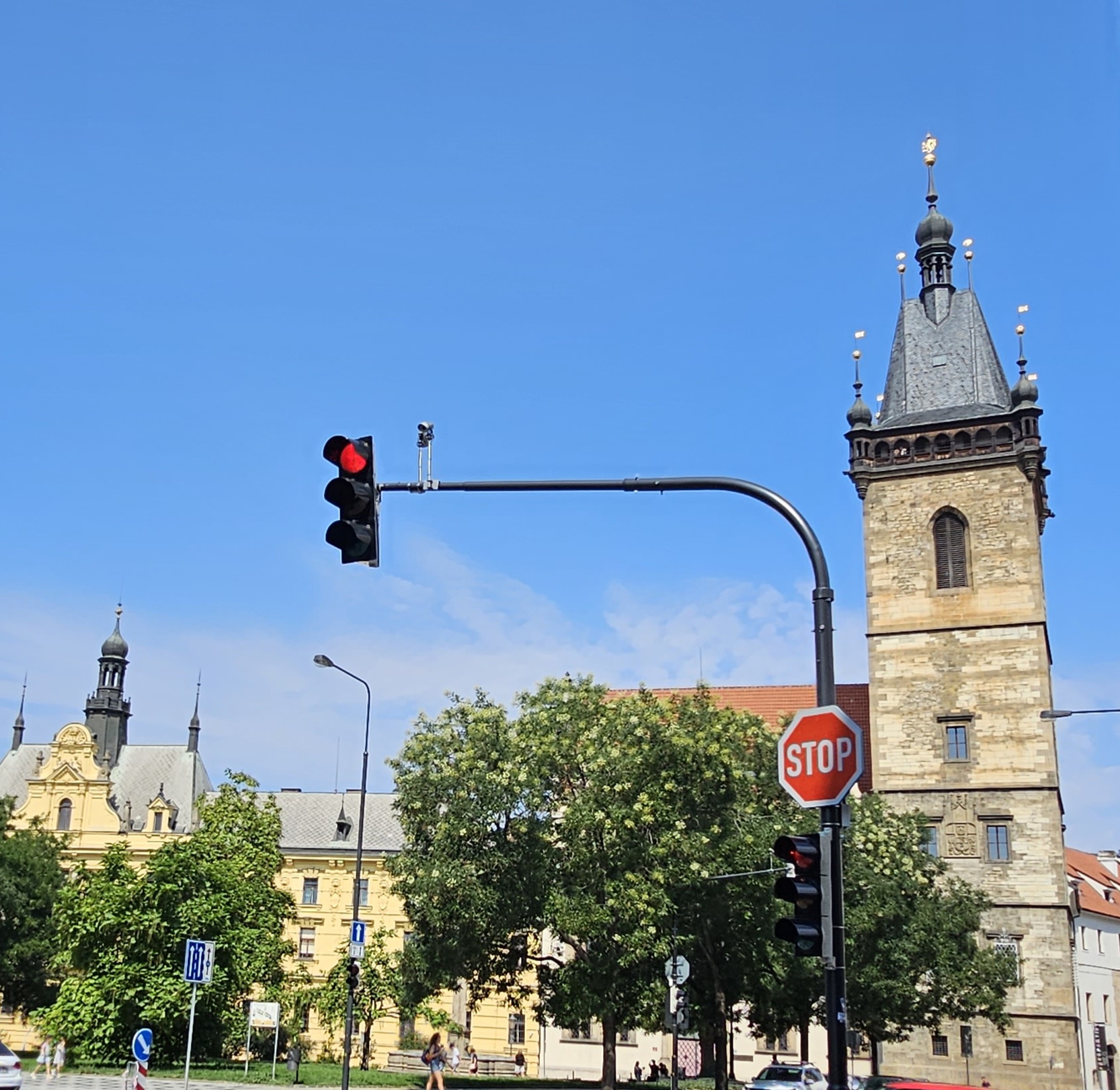 The New Town Hall in Prague, where the first Defenestration took place in 1419. A tower with three windows can be seen across road. In front of the town hall are some trees. There is a traffic light above the trees.