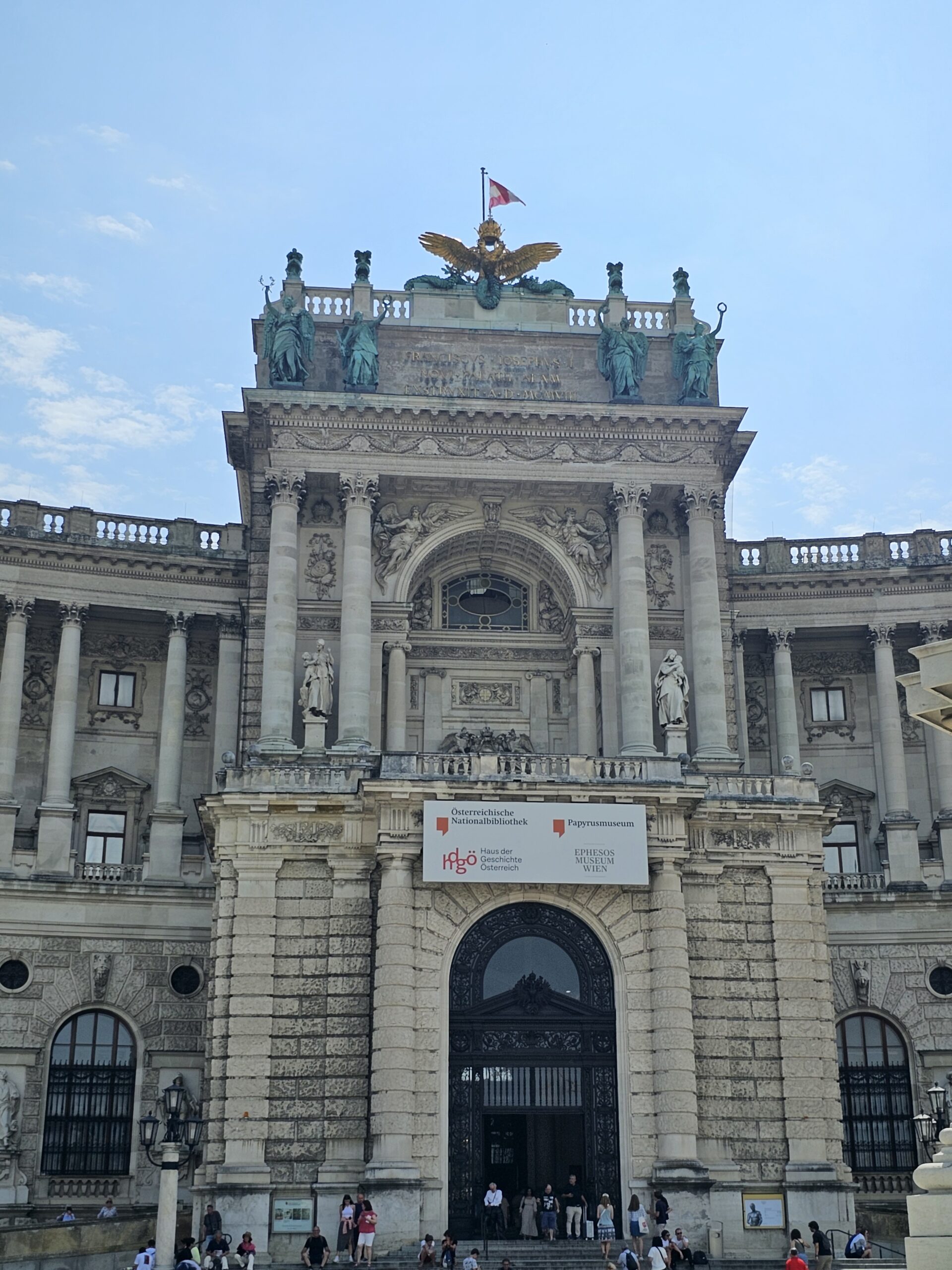 Front facade of Hofburg Palace. The door is surrounded by columns with green statues at the top. The roof has a golden eagle with wings spread. Image by 360onhistory.com