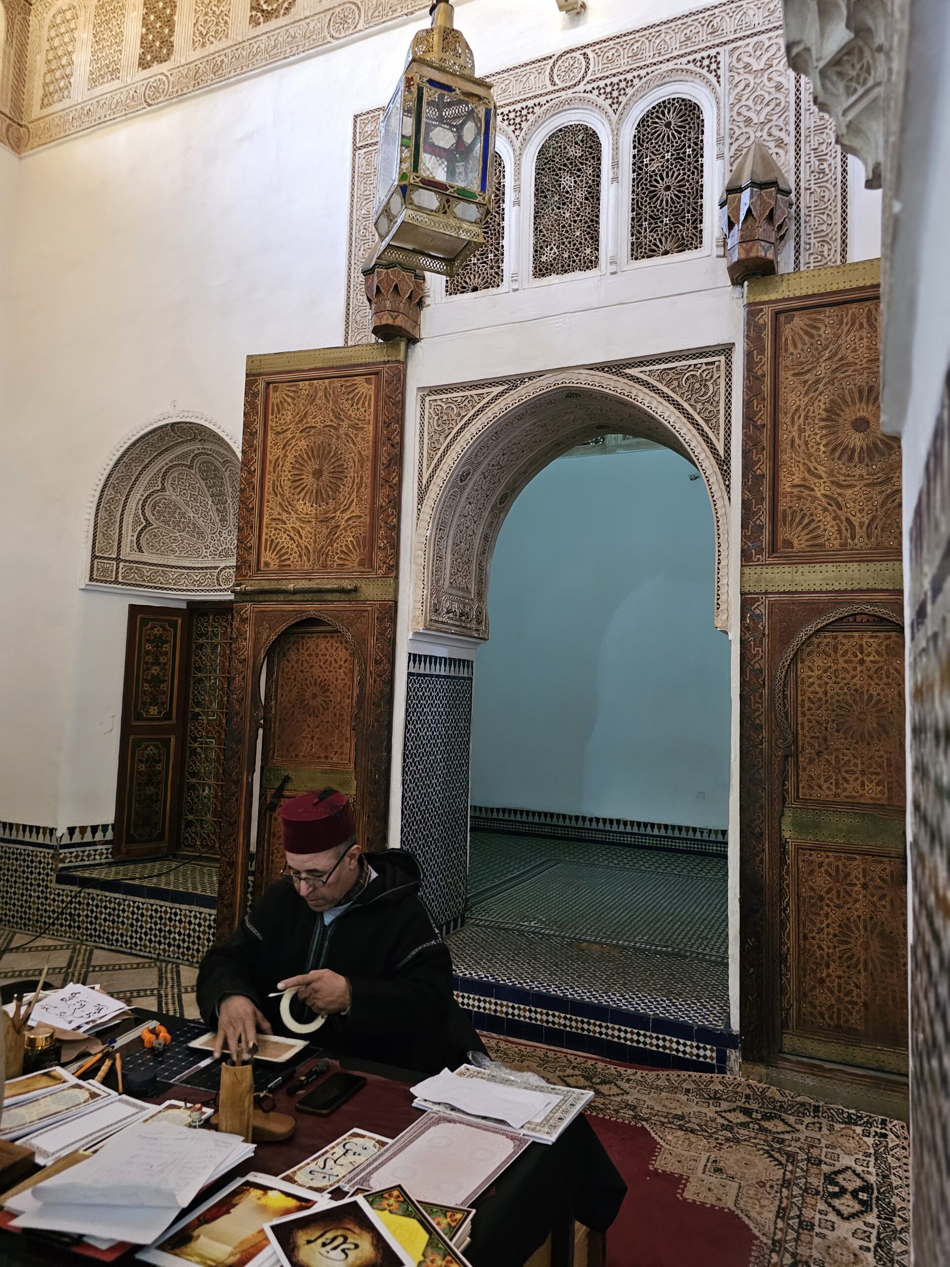 A scribe sits on the floor in front of a doorway at Bahia Palace, Marrakesh. He has books and papers in front of him. Image by 360onhistory.com