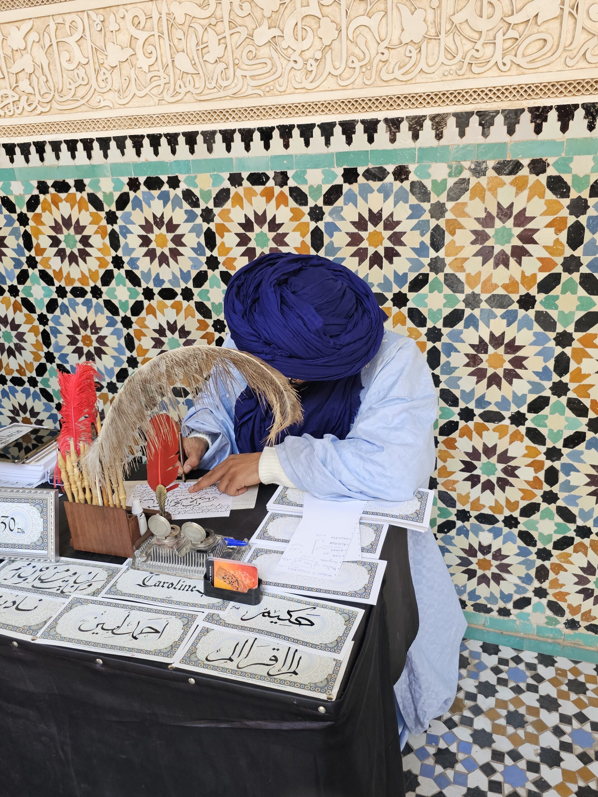 A khateeb (scribe) in a blue turban and white clothes sitting at a table and doing calligraphy. Image by 360