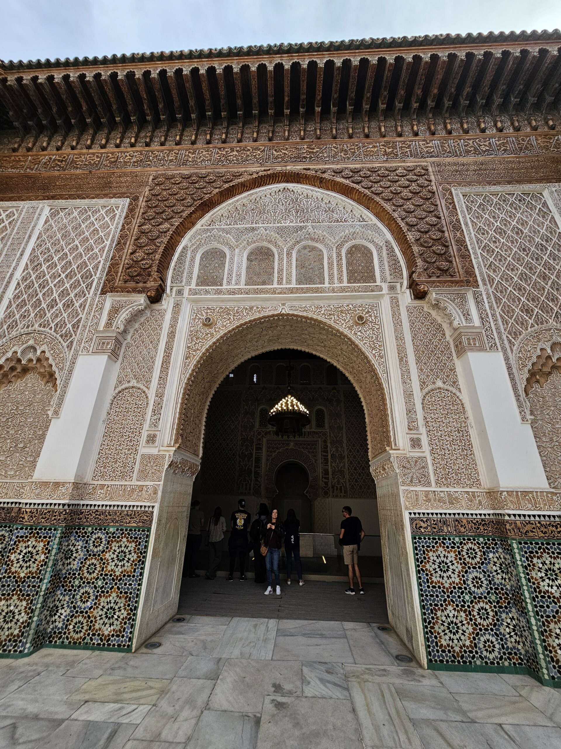 An ornate wooden door with beautifully carved surrounding walls which have colourful Islamic tiles at the lower half at Ben Youssef Madrasa, Marrakesh. Image by 360onhistory.com