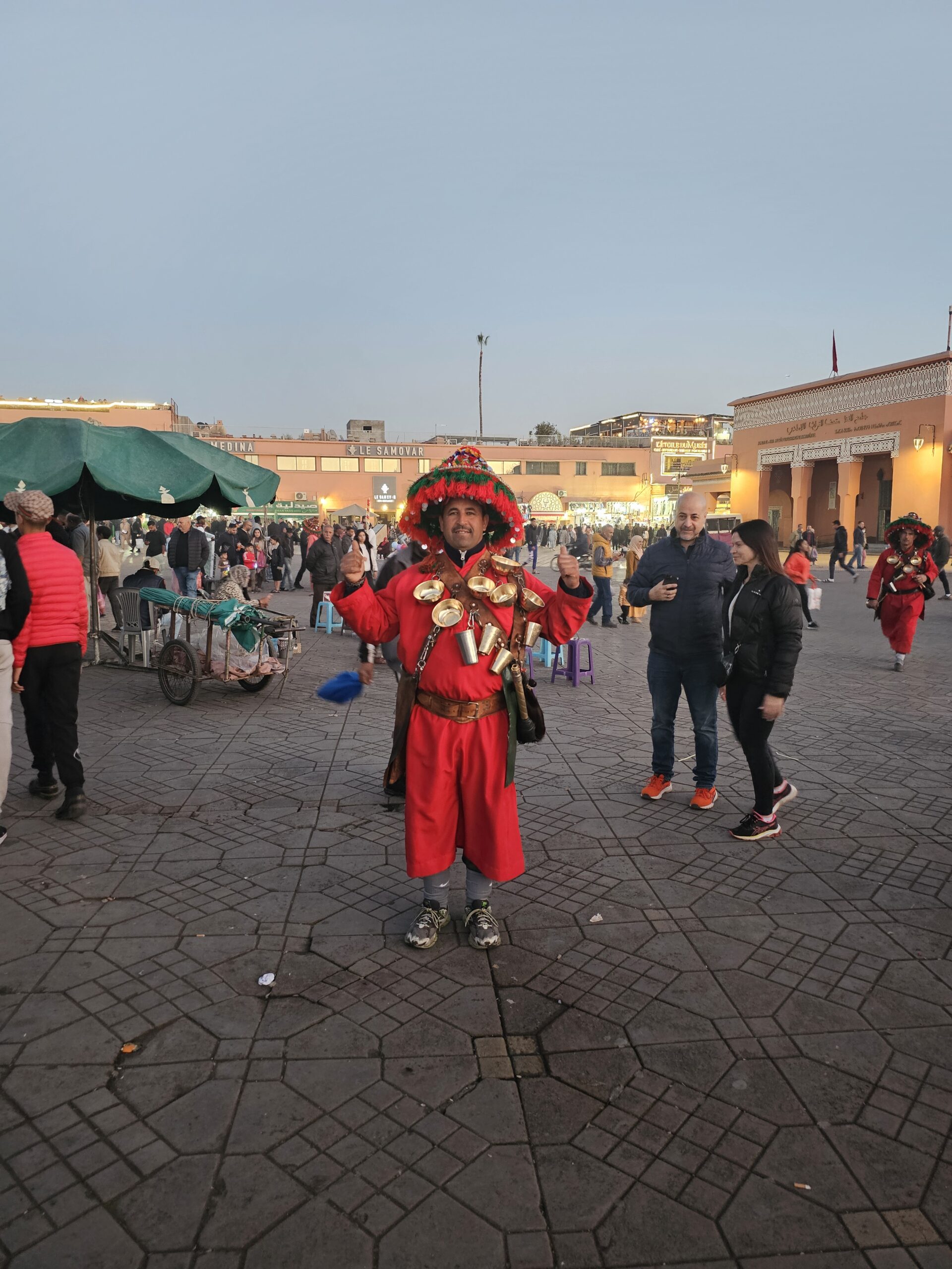 Traditional water seller or "Gharrib," in Jemaa El Fnaa, Marrakesh. He is wearing bright red clothes and hat and has brass water utensils hung around him. Image by 360onhistory.com